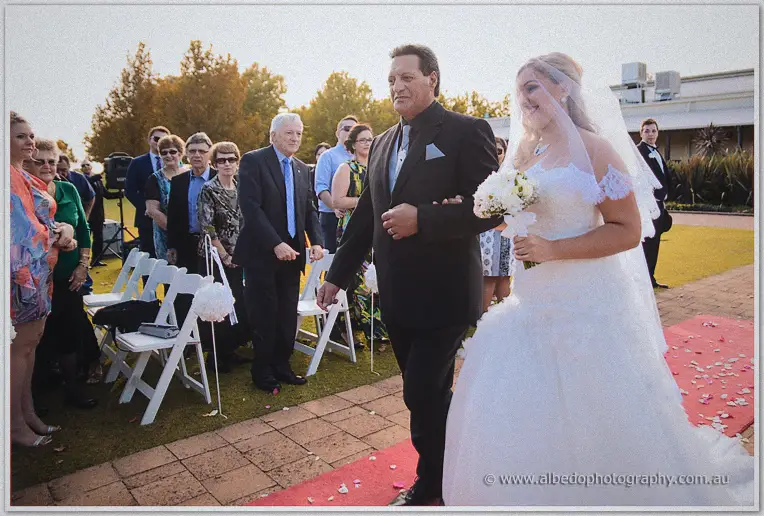 Jasmine and her father passing rows of seated guests on the red carpet aisle in soft late-afternoon light