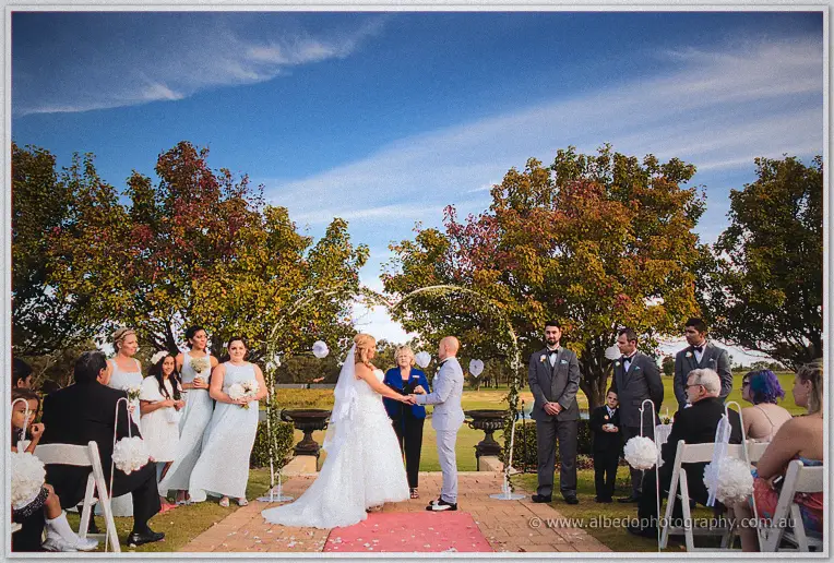 Jasmine and Michael joining hands under twin arches, bridal party and guests beneath a big blue sky