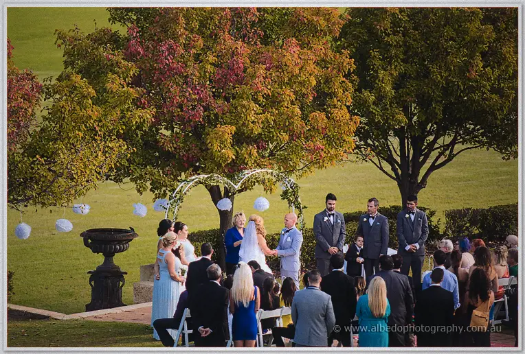 Jasmine and Michael exchanging vows beneath the white arbour as celebrant and guests watch on