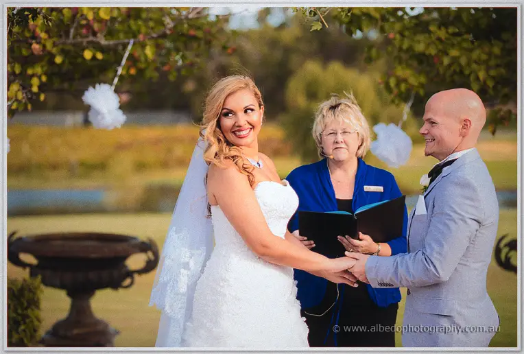 Jasmine and Michael beaming as they hold hands at the altar, celebrant reading between them by the water
