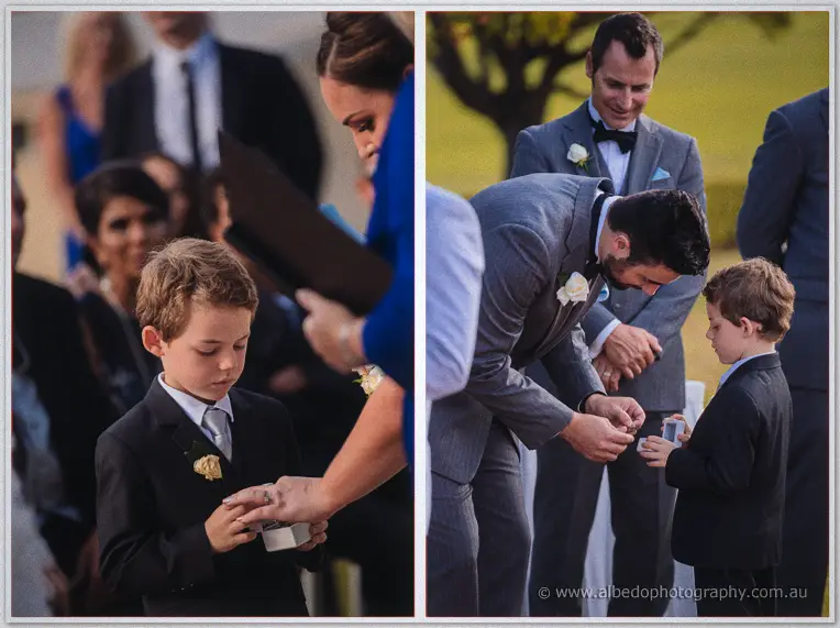 A young page boy in a black suit hands the wedding rings to Michael beneath the trees.