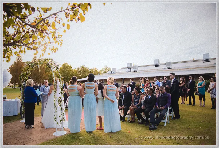 Wide view of the outdoor ceremony on the winery lawn with bridesmaids in pale blue and seated guests.