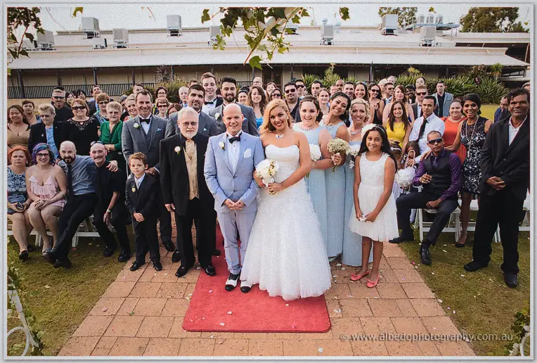 The whole wedding party gathers on the red-carpet aisle in front of the winery building.