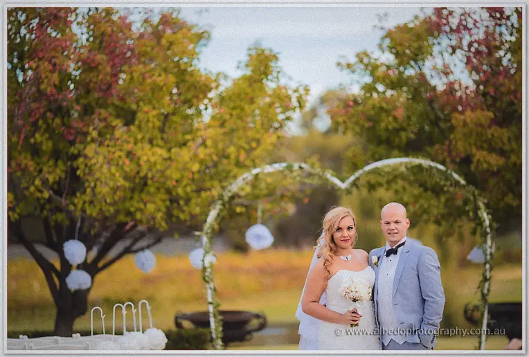 The bride and groom stand arm in arm beneath autumn trees and white paper pom-poms.