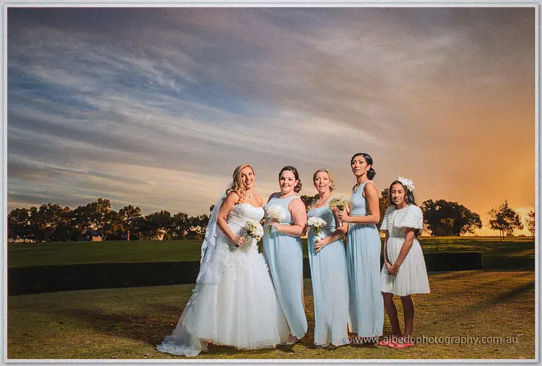 Bridesmaids in pale blue join the bride beneath a dramatic sunset sky on the lawn.