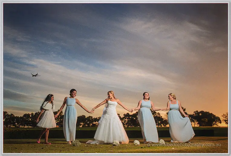 The bride and her bridesmaids hold hands and laugh under a sunset sky with a plane overhead.