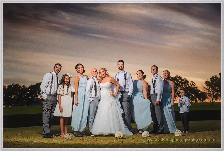 Full bridal party in blue gowns and braces poses under streaky dusk clouds.