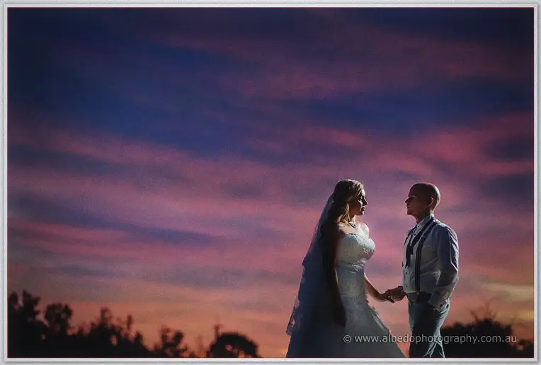 The newlyweds face each other hand in hand beneath a vast violet and coral sunset.