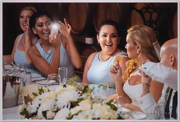 Bridesmaids laugh as one films on her phone beside the bride at the bridal table.