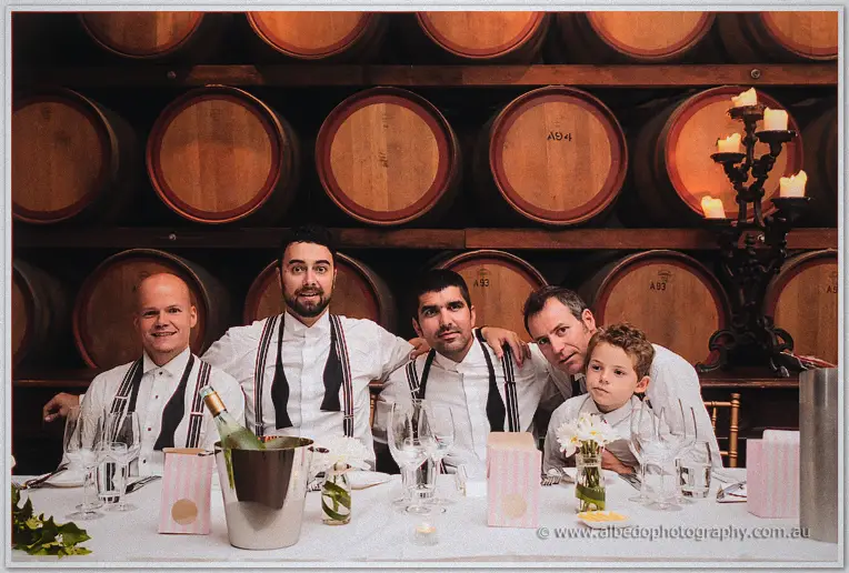 Groomsmen in braces and a page boy line the table before stacked wine barrels.