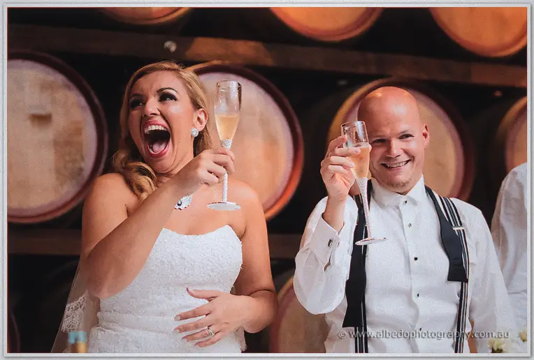 Jasmine and Michael cheer with raised champagne flutes in front of the barrels.