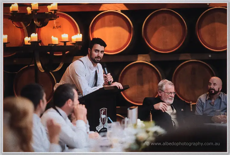 A best man leans on the podium mid-speech before the glowing barrel wall.