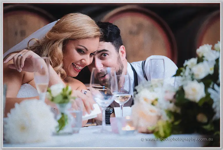 Jasmine laughs with a groomsman at the candlelit bridal table, wine barrels glowing behind the glassware.