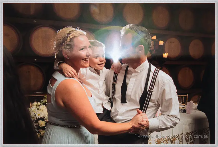 A flower-crowned boy between two beaming adults joins a family dance beneath the wine barrels.
