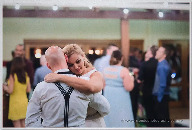 Jasmine wraps her arms around a guest in suspenders, eyes closed mid slow dance in the crowded room.