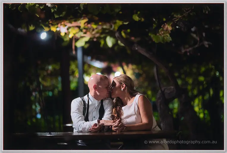 Jasmine and Michael lean across a timber table for a kiss beneath the vine-covered pergola at night.
