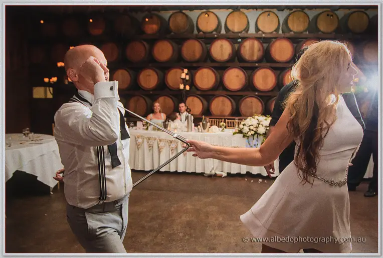 Jasmine playfully hooks Michael's suspenders with a cane as the bridal table watches and laughs.