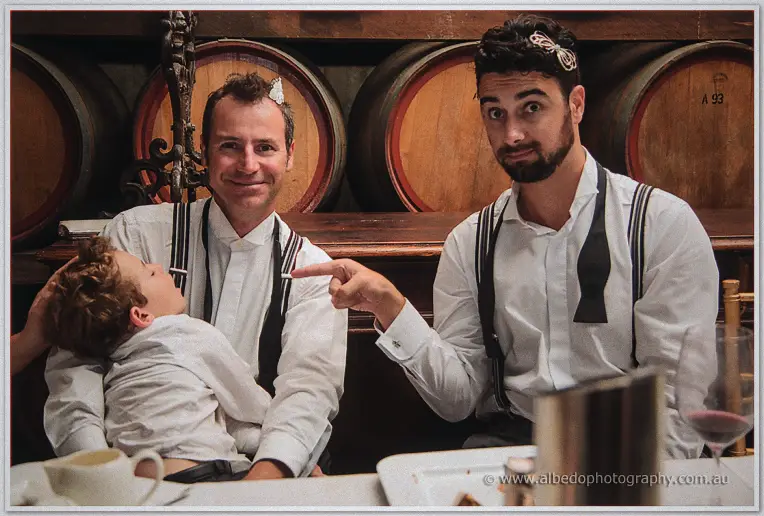 Two groomsmen in suspenders clown for the camera, one cradling a sleepy page boy by the barrels.