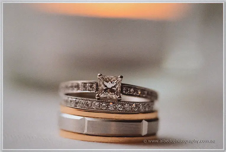 Jasmine's solitaire ring rests atop Michael's bands, candlelight blazing soft gold behind the macro shot.