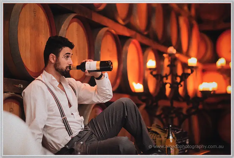 A groomsman swigs straight from a wine bottle, lounging among candlelit barrels in the cellar.
