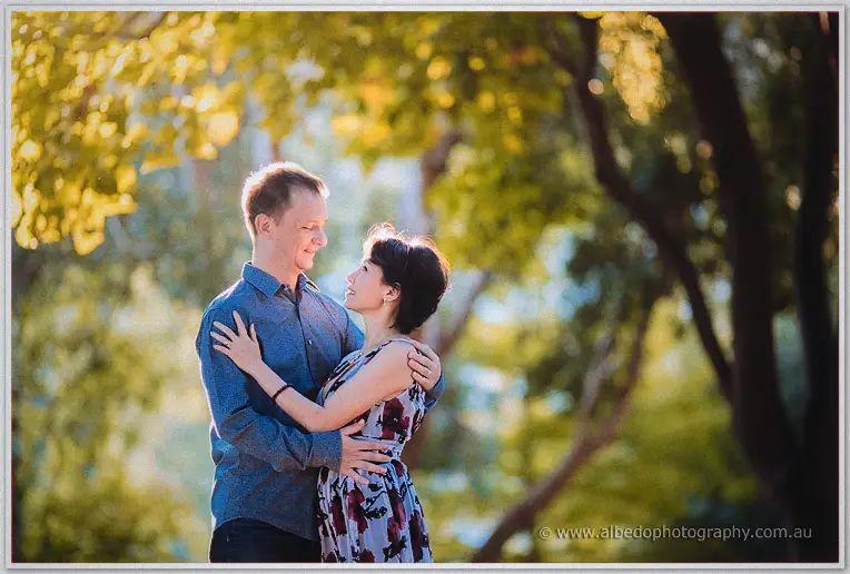 Loan wraps her arms around Alex under sunlit gum trees, both laughing in warm afternoon light