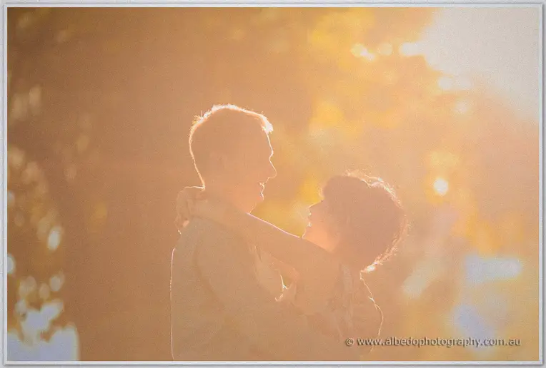 Loan and Alex dissolve into sun flare as they hug close under backlit autumn trees