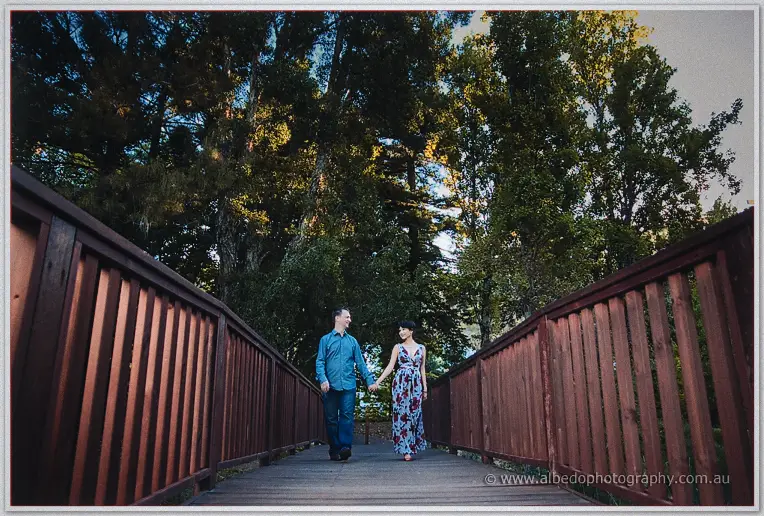 Loan and Alex walk hand in hand across a red timber footbridge beneath tall sunlit pines