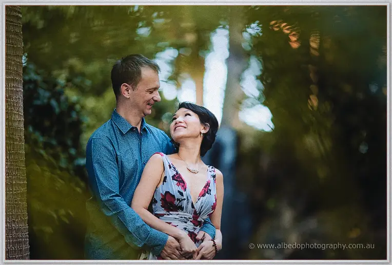 Loan leans back against Alex and gazes up at him amid soft green park foliage