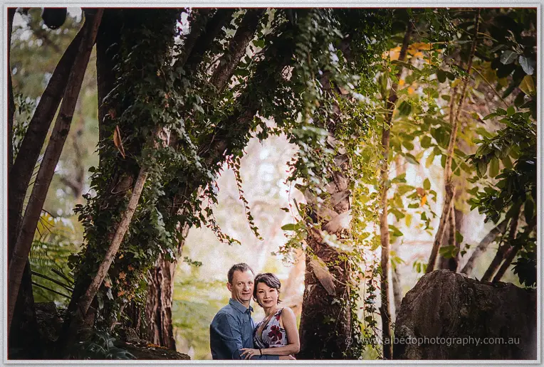 Loan and Alex stand close beneath a vine-draped tree arch as evening light glows behind them