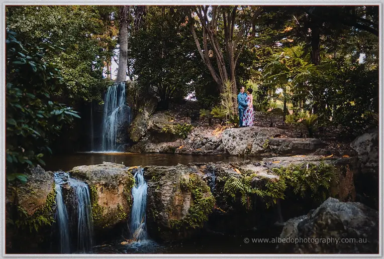 Wide view of Loan and Alex posing on mossy rocks beside waterfalls in a lush garden