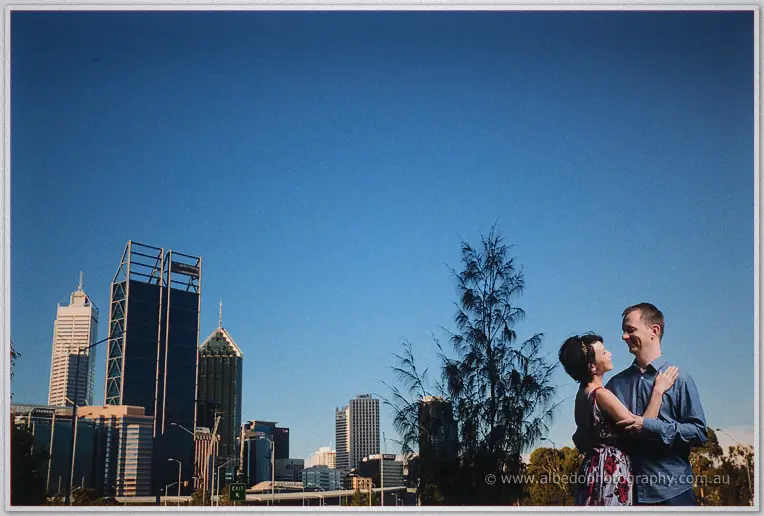 Loan rests her hands on Alex's chest as they laugh beside city towers and native trees