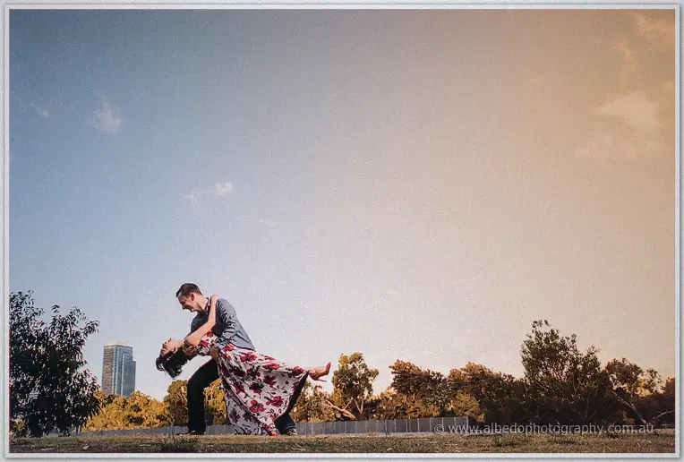 Alex dips Loan low across a grassy park lawn with the city rising behind them at dusk