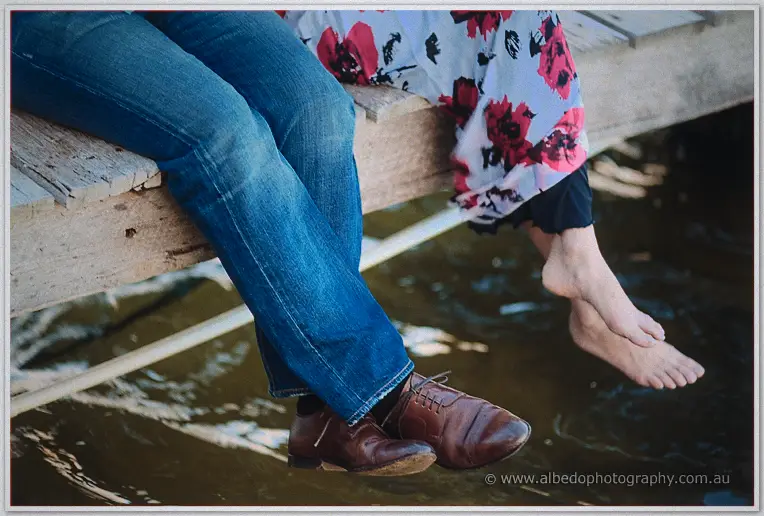 Close-up of Alex's brown dress shoes and Loan's bare feet dangling from the jetty above the river