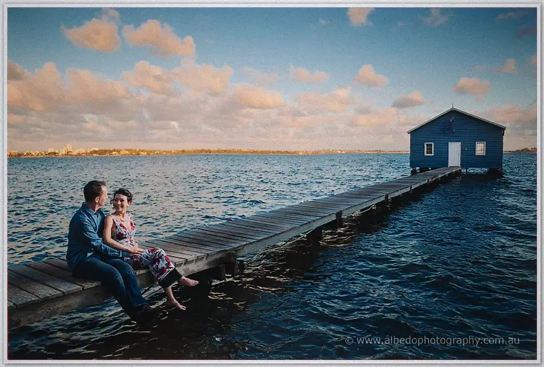 Loan and Alex laugh while sitting on the long jetty leading to the blue boat house at dusk