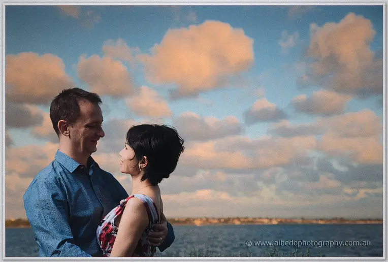 Loan and Alex smile at each other by the river under a sky of pink sunset clouds