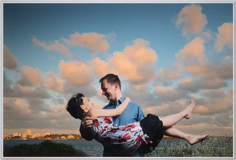 Alex carries laughing Loan in his arms against the evening sky above the water