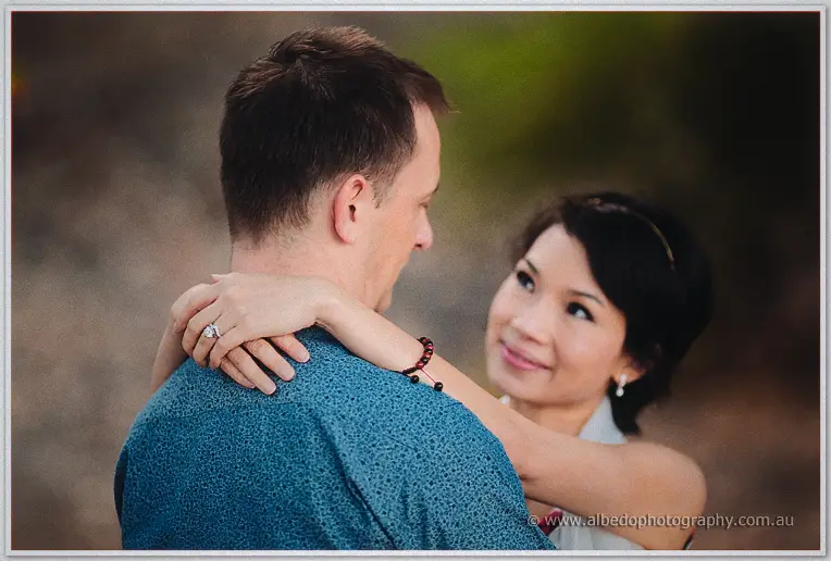 Loan gazes up at Alex with clasped hands on his shoulder, her ring catching the light