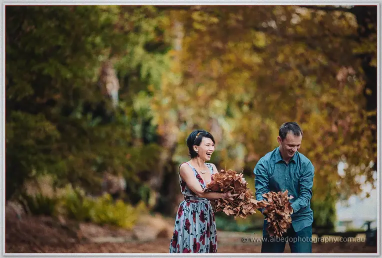 Loan and Alex laugh as they gather armfuls of dry autumn leaves beneath golden trees