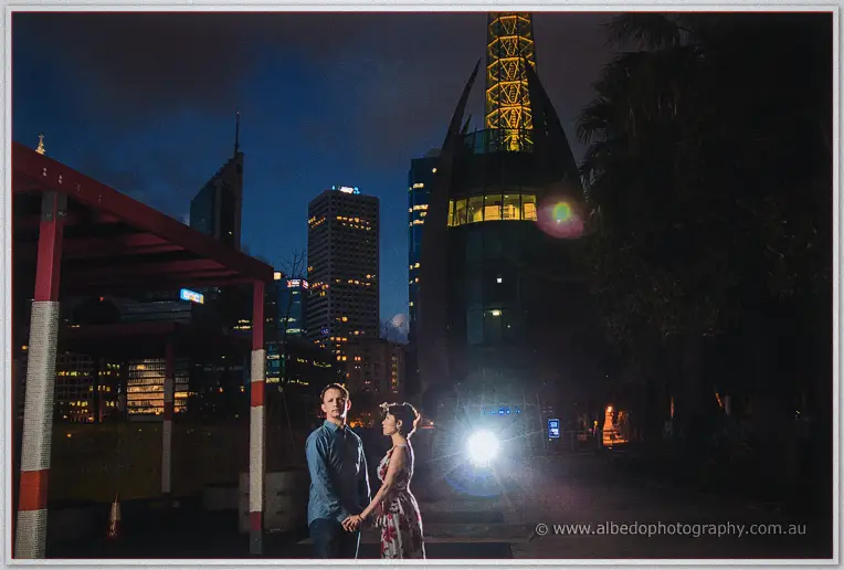 Loan and Alex stand hand in hand before the lit bell tower and city towers after dark