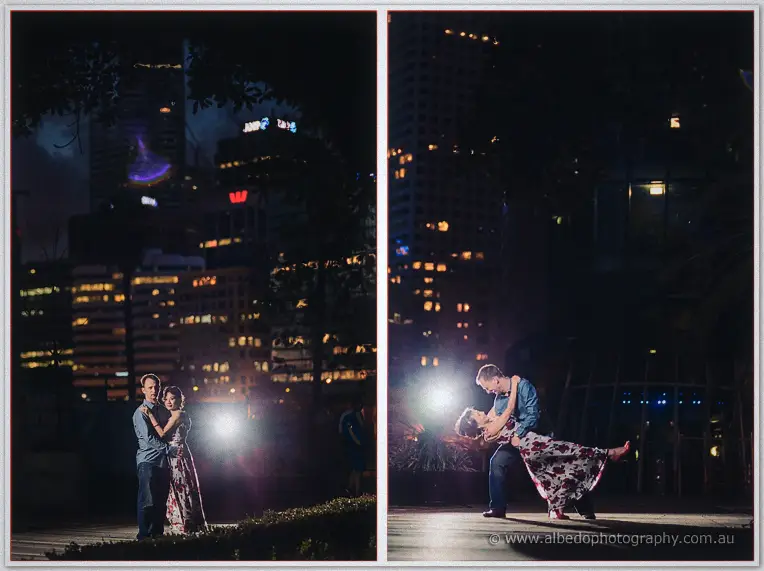 Alex dips Loan under city lights as a bright flash glows between the night towers