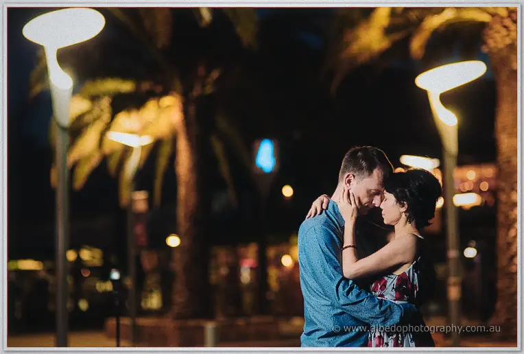 Loan holds Alex's face as the pair embrace close beneath glowing palm trees at night