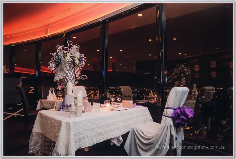 A lace-draped sweetheart table with tall candles and purple sashed chairs glows by dark windows