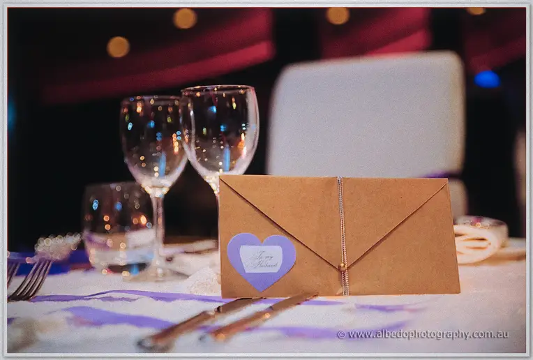 A kraft envelope with a purple heart and wine glasses rests on the lace reception table