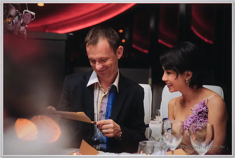 Alex laughs reading a card at the table while Loan beams beside him under red drapery