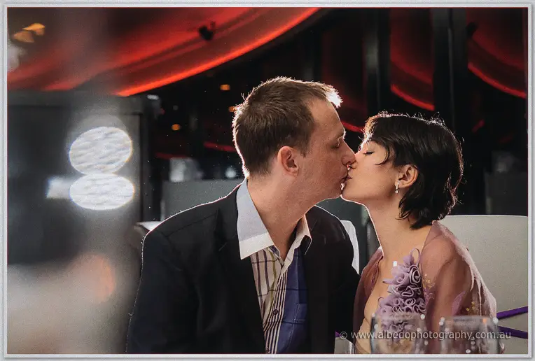 Alex and Loan share a kiss at their candlelit table beneath sweeping red ceiling drapes