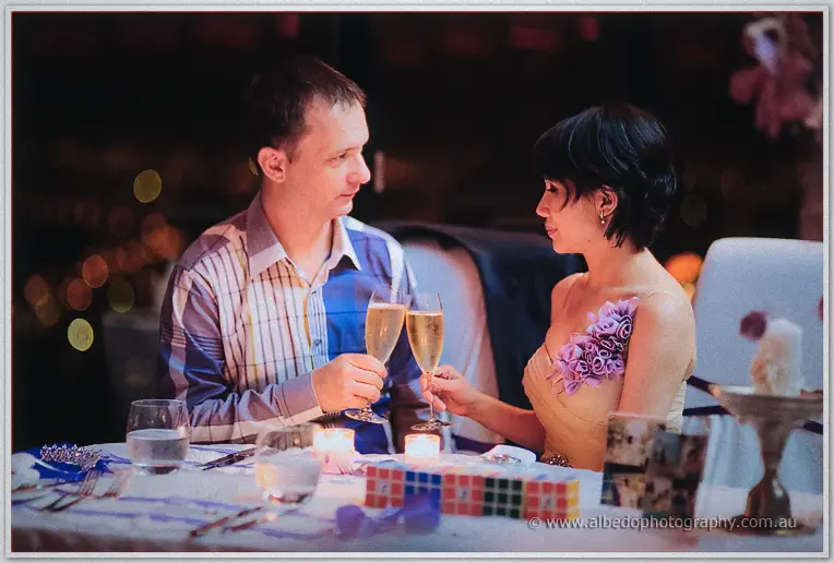 Alex and Loan toast with champagne flutes across their table as city lights blur outside