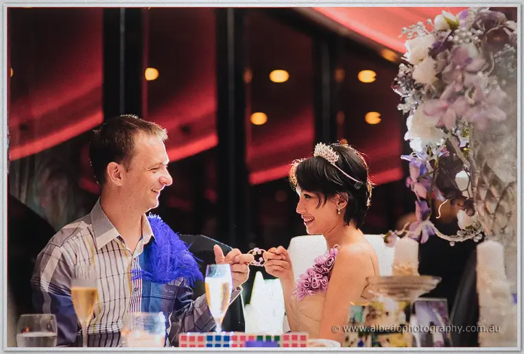 Loan and Alex clink hands across an anniversary dinner table, her tiara and purple blooms glowing warmly.
