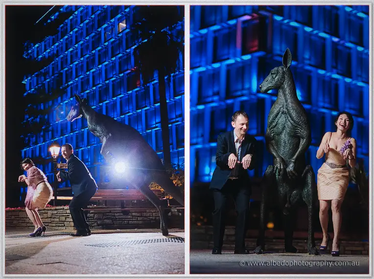 Loan and Alex hop beside bronze kangaroo statues under Perth's blue-lit Council House towers at night.