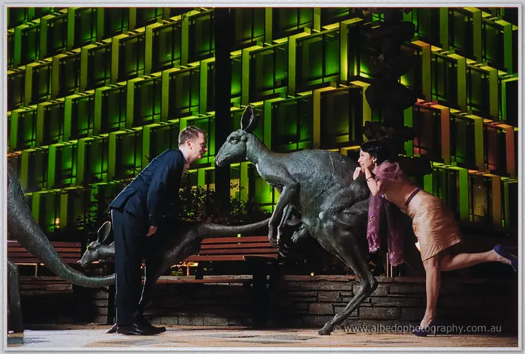 Alex leans nose to nose with a bronze kangaroo while Loan crouches behind it laughing under green city lights.