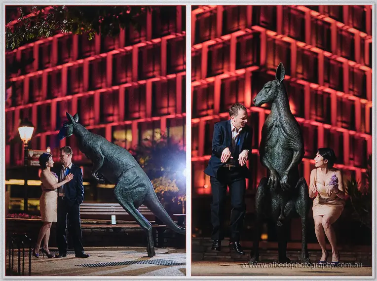 Loan and Alex embrace under a street lamp, then leap around a kangaroo statue beneath red-lit towers.